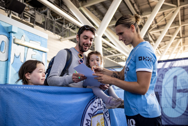 A Manchester City player in a blue jersey signs an autograph for a man holding a child, with another child beside them, at a stadium with Manchester City decorations.