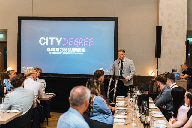 A speaker is at a podium during the 2025 graduation ceremony for the City Degree program in Manchester City Community Football Coaching, with attendees seated at dining tables.