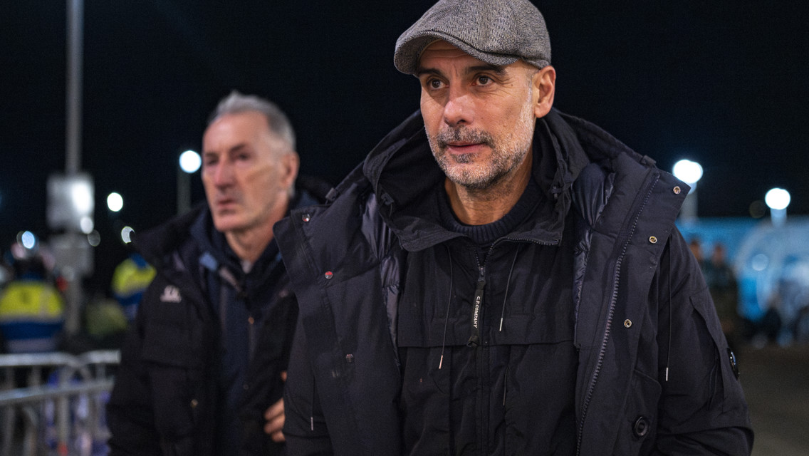 Two individuals with blurred faces wearing dark coats and hats at a night event with lights in the background.