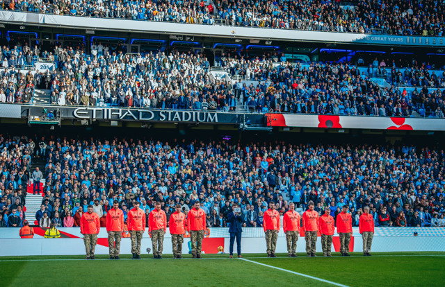 Manchester City fans at the Etihad Stadium during a Remembrance Sunday observance, featuring individuals wearing commemorative attire and standing in line.
