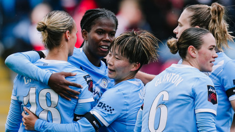 Manchester City Women's team players celebrating a goal in light blue jerseys with the club's logo visible. Players are embracing, with visible player numbers like 18 and 6.
