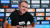 Person at a press conference wearing a black shirt, seated at a table with microphones, drinks, and various sponsor logos in the background.