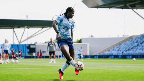 A soccer player wearing a Manchester City women's team kit controls the ball on a pitch. The background shows a stadium with spectator stands.