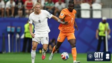 Two soccer players in action on the field, one in a white jersey and the other in an orange jersey, during a match at the City World Cup.