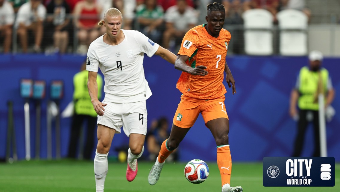 Two soccer players in action on the field, one in a white jersey and the other in an orange jersey, during a match at the City World Cup.