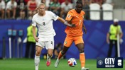 Two soccer players in action on the field, one in a white jersey and the other in an orange jersey, during a match at the City World Cup.
