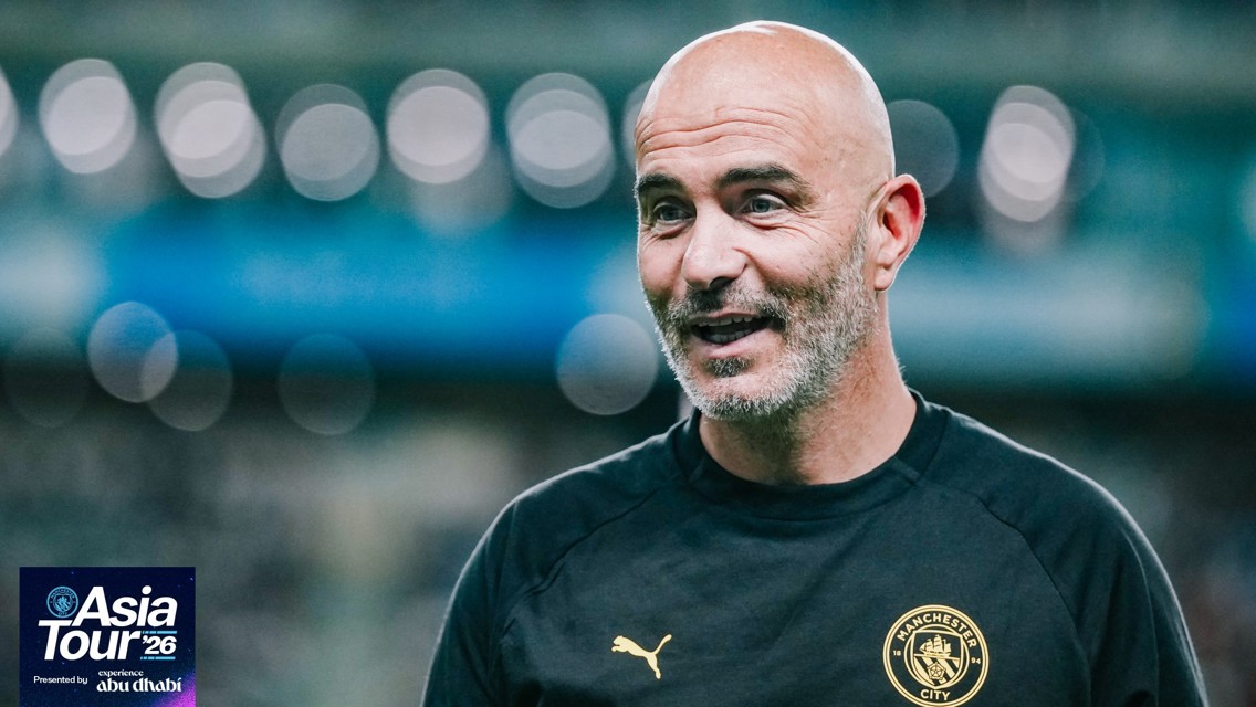 A Manchester City coach wearing a club shirt with a Puma logo and the team's crest, smiling during the Asia Tour 2026. Background is blurred with circular light patterns. The tour is presented by Experience Abu Dhabi.