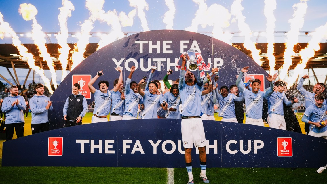 Manchester City Youth Team celebrating their FA Youth Cup victory with fireworks in the background.