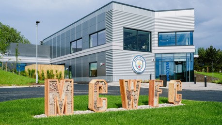 Modern building with Manchester City crest and large letters 'MCWFC' made of wooden frames filled with materials on a grassy area.