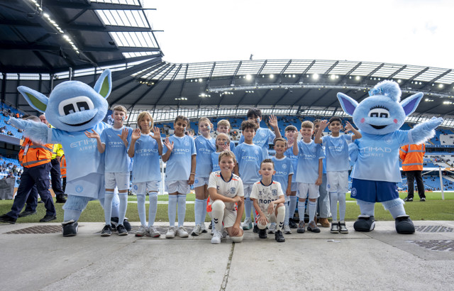 A group of children wearing Manchester City jerseys pose with club mascots Moonbeam and Moonchester at the Etihad Stadium.