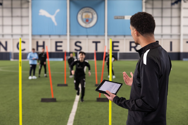 A coach holding a tablet observes players running through training poles on an indoor field with Manchester City branding.