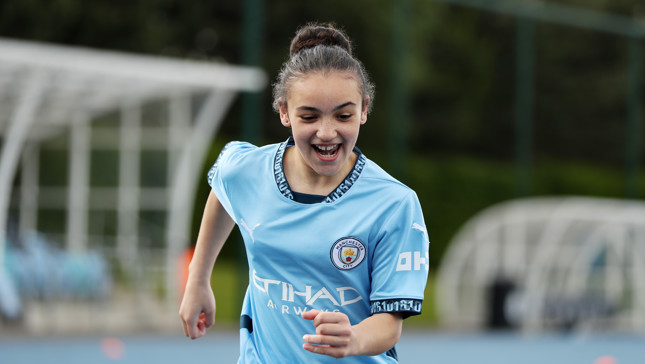 A football player wearing a Manchester City blue jersey is captured in action, with the blurred background of a soccer field.