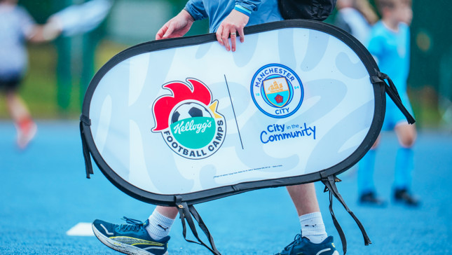 Person carries a sign promoting Kellogg's Football Camps and City in the Community by Manchester City. Logos are on the sign against a blue playing field backdrop. Event involves football activities for children.