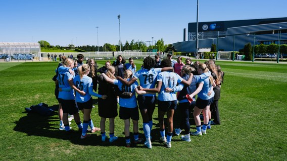 A group of women soccer players in light blue jerseys form a huddle on a grassy field. A building in the background displays the Manchester City logo.