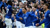 Everton players wearing blue jerseys celebrate together during a football match, surrounded by cheering fans.