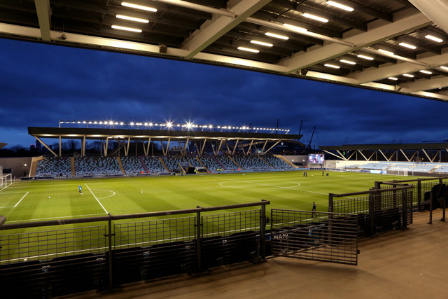 The photo captures an evening view of Academy Stadium, featuring a well-lit football field and empty spectator stands.