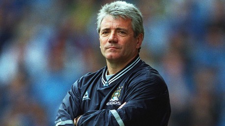 A man with gray hair wearing a Manchester City jacket, standing with arms crossed.
