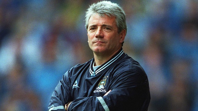 A man with gray hair wearing a Manchester City jacket, standing with arms crossed.