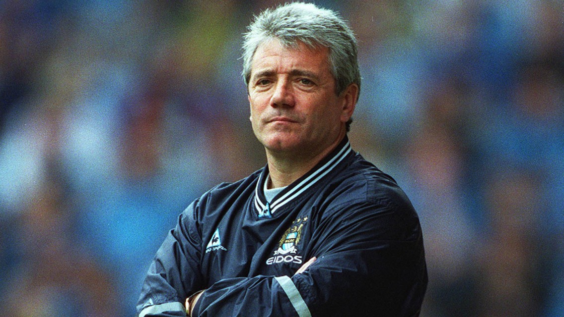 A man with gray hair wearing a Manchester City jacket, standing with arms crossed.