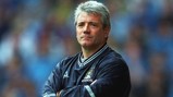 A man with gray hair wearing a Manchester City jacket, standing with arms crossed.