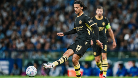 A Manchester City player in a black and yellow kit is kicking a soccer ball during a match. Another player in the background is wearing the same kit.