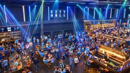 A bustling indoor event at City Hall with attendees dressed in light blue clothing and socializing around tables and bars under vibrant lighting.