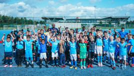 Group of children and teenagers in Manchester City football kits on a football field raising their fists in celebration with a stadium in the background.