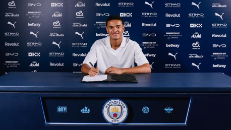 A person sitting at a table signing a contract at Manchester City. The background features numerous sponsor logos.