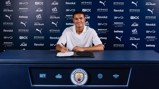 A person sitting at a table signing a contract at Manchester City. The background features numerous sponsor logos.