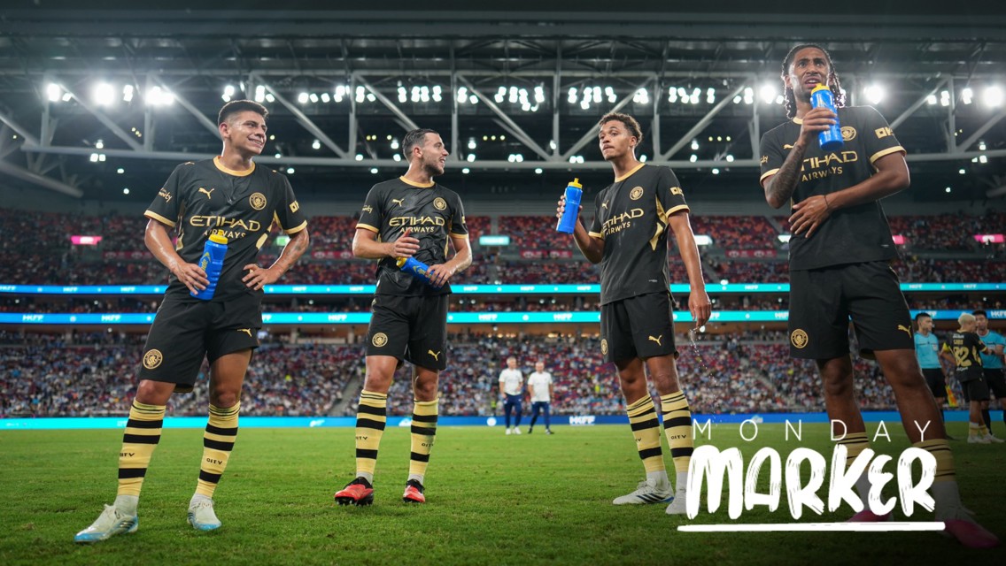 Four Manchester City football players in black and gold kits are holding blue water bottles on a football field, with stadium lights and crowd in the background. Text reads 'Monday Marker'.