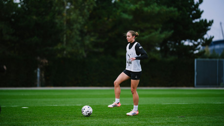 A woman in sportswear practicing soccer on a grassy field with a ball nearby. She wears a white bib with "Puma" branding.