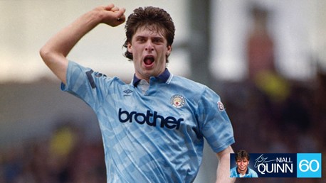 A football player in a vintage Manchester City kit celebrates with a clenched fist. The shirt features 'brother' as the sponsor and the Manchester City crest on the chest.