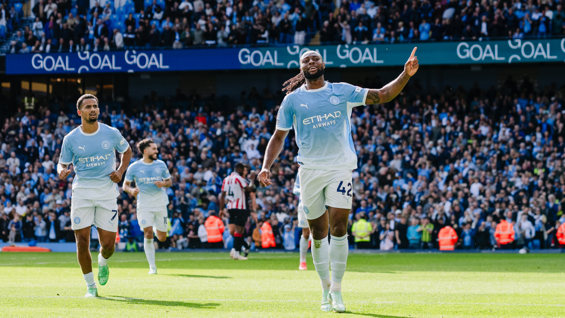 Manchester City players celebrating a goal at Etihad Stadium. The players are wearing light blue uniforms as one leads with a gesture amidst a cheering crowd.