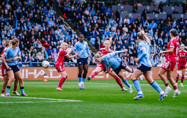 Women's soccer match with players from teams in light blue and red jerseys competing for the ball in front of a crowded stadium.
