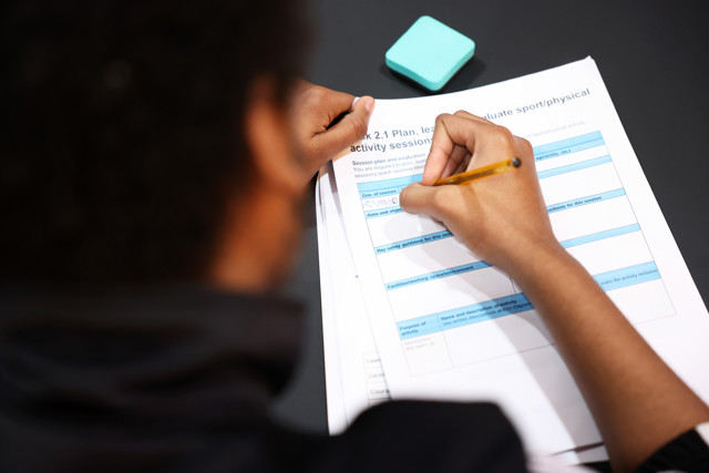 A person writing on a worksheet about planning and evaluating sport/physical activity sessions. The page includes sections for session details, aims, safety guidance, and facilities.