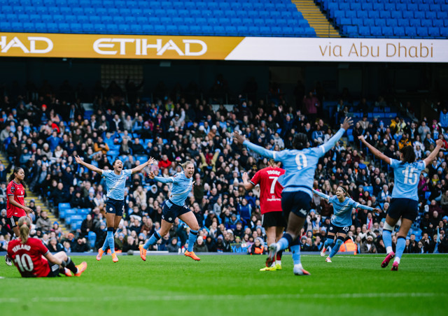 Football players in light blue jerseys celebrate on the field while opposing players in red observe. The background shows a crowd and 'Etihad' signage at a stadium.