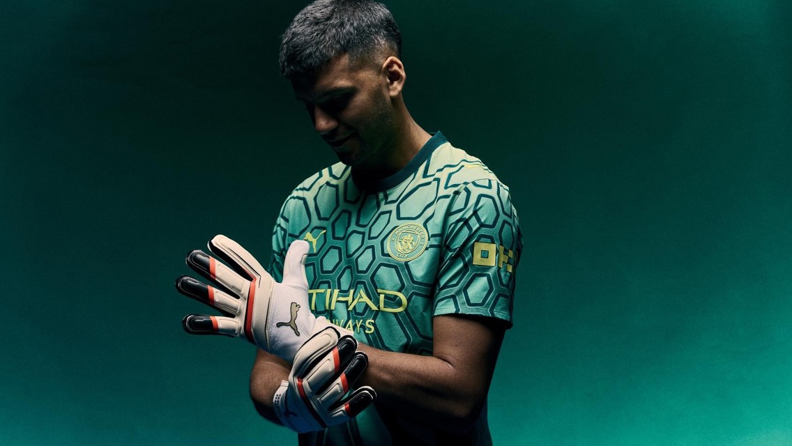 A person wearing a Manchester City goalkeeper kit, adjusting their gloves. The shirt has hexagonal patterns and the Manchester City crest is visible.