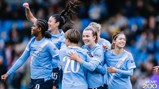 A group of female football players in light blue jerseys celebrate together on the field. Player number 20 is being hugged by teammates, and they all have smiles on their faces.