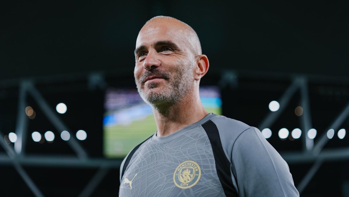 A bald man wearing a Manchester City shirt with a logo stands in a stadium with blurred lights in the background.