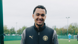A man smiling while wearing a Manchester City jacket standing in front of a sports field.