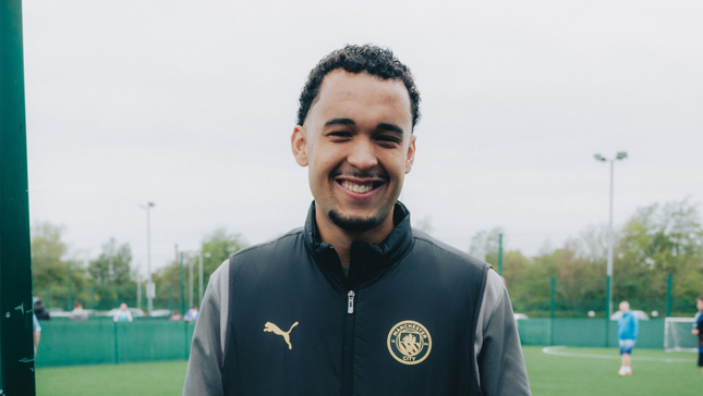 A man smiling while wearing a Manchester City jacket standing in front of a sports field.