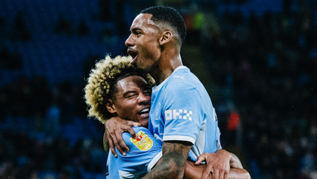 Two soccer players in light blue jerseys embrace in celebration during a match.