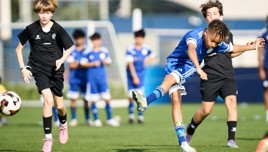 Youth soccer players in action during a match, with one player in blue kicking the ball while others in black run nearby.