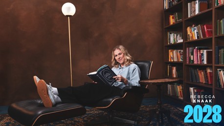 A person is seated on a black leather chair reading a book in a cozy library setting with shelves full of books in the background. In the bottom right, 'Rebecca Knaak 2028' is written in blue.