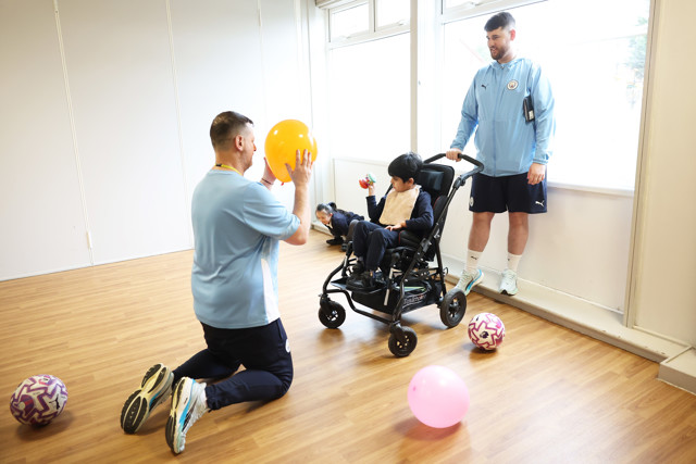 Two adults and a child in a wheelchair indoors with footballs and balloons. The adults wear light blue shirts with a Man City logo.