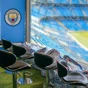 Interior view of Etihad Stadium showing Manchester City logo, seating area with black chairs, and pitch in the background.