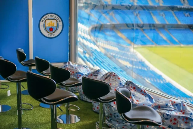 Interior view of Etihad Stadium showing Manchester City logo, seating area with black chairs, and pitch in the background.