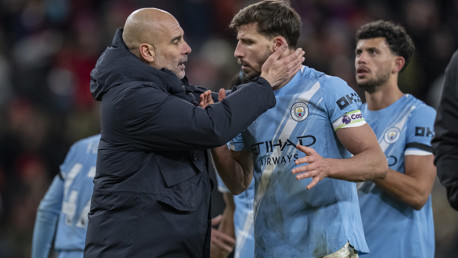 Manchester City players interacting on the field with blurred faces; one wearing the team kit and the other in a jacket.