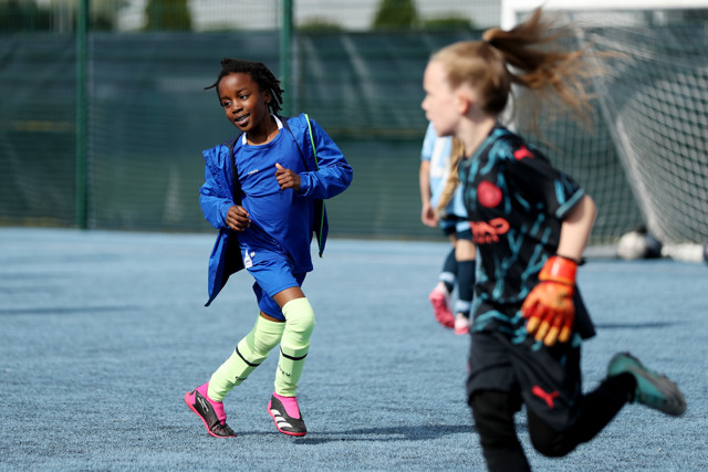 Two young children playing soccer on an outdoor pitch. One child in blue running with the ball while another child in black, wearing goalkeeper gloves, is running towards the goalpost.