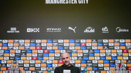 A press conference setup with Manchester City branding and sponsor logos on the background. Bottles of beverage are placed on the desk in front of empty chairs.